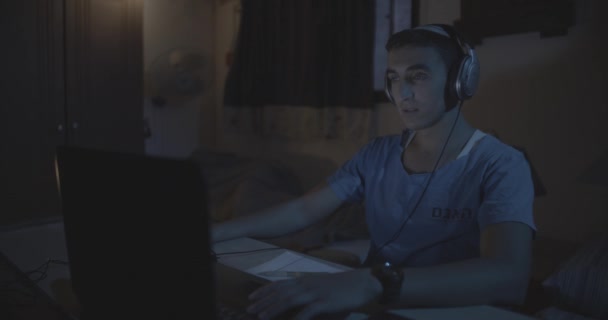 파란색 티셔츠를 남자가 어두운 탁자에 헤드폰을 컴퓨터 — 비디오