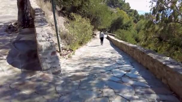 マラガのジブラルファロ城から天然石で覆われた小さな道を歩く女性 マラガの人気の観光スポット 丘の下の街の美しい景色 — ストック動画