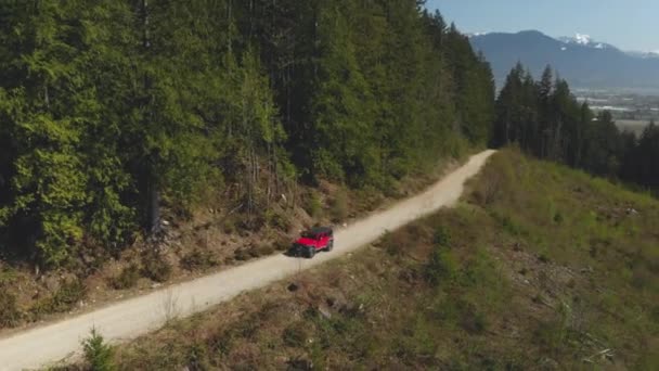 未舗装の伐採道路に接近し バックグラウンドで密な緑の森の景色を通過する道路車両のオフレッドジープSuvトラックがパンを通過するように追跡を見下ろす上の空中が安定した — ストック動画