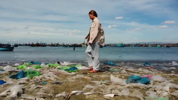 看到白人女孩在塑料袋上走在海岸上 背景中的渔船 — 图库视频影像