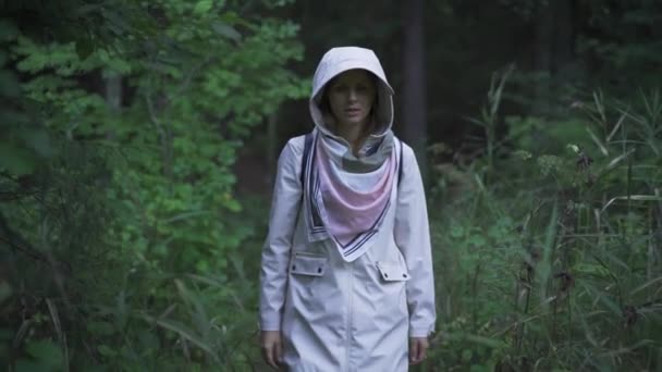 穿着白色雨衣在森林里行走的女人 她正朝摄像机前走去 从树阴下进入光带时摘下了她的头罩 — 图库视频影像