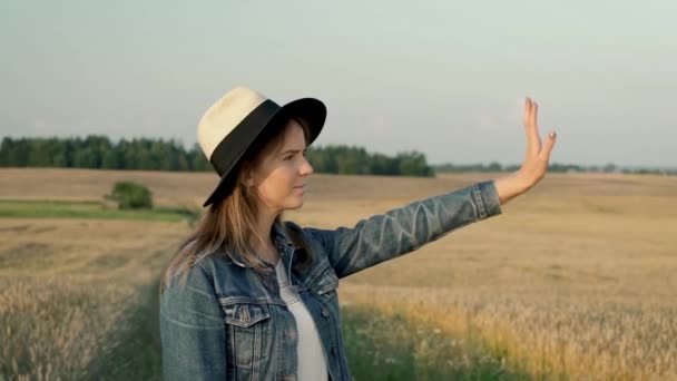 女の子は空中で手を振って誰かに挨拶をしています 女性はデニムジャケットと夏のスタイルの帽子をかぶっています 彼女は国中を旅行する休日の幸せな旅行者のように見えます — ストック動画