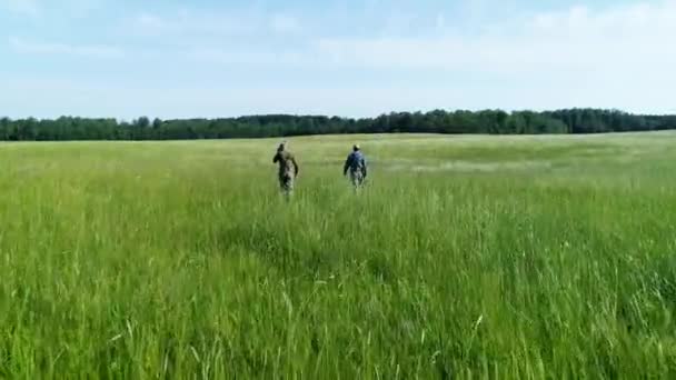 在夏天的白天 你可以看到两个人在绿色的草地上散步 照相机在飞行 从后面跟着朋友们 而他们正与森林一起向地平线飞去 — 图库视频影像