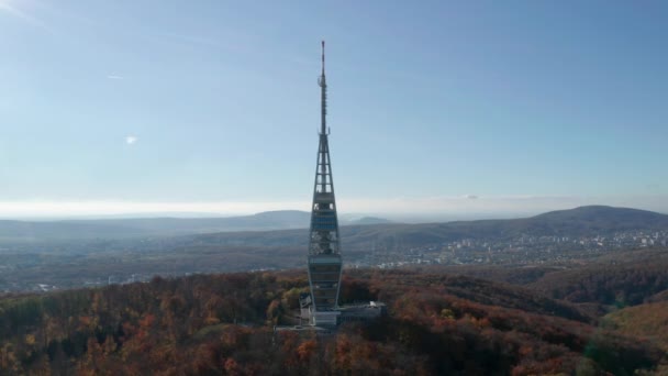 Aerial orbit view of a TV tower with a large antenna called Kamzik in Bratislava, Slovakia. Autumn backlit scenery with colourful trees around the tower and blue sky. Cityscape is in the background.