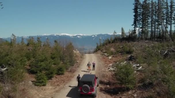 三个人从停在山顶上的一条土路上的吉普车旁边走过 在明亮的蓝天空中 他们可以看到覆盖着山谷雪的山峰 — 图库视频影像