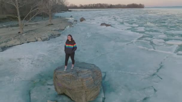 在冰冻湖中的岩石上穿着骄傲色彩外套的年轻女人 — 图库视频影像