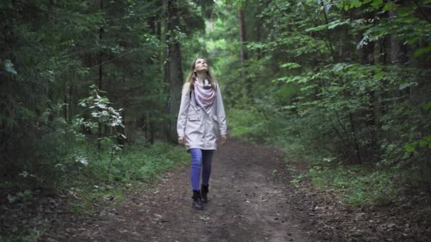 青いジーンズと白いレインコートを着た森の中を歩く女性 彼女は緑の植物 木に囲まれた森のどこかの遠隔地の野生の森林経路に立っています — ストック動画