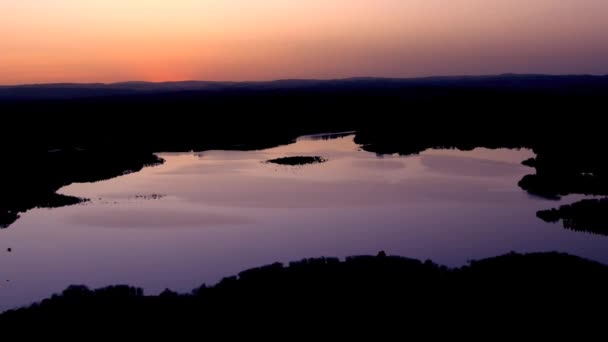 穏やかな滑らかな湖の上には 森に囲まれたオレンジの夕日の空に紫色の反射光があります ドリー バック — ストック動画