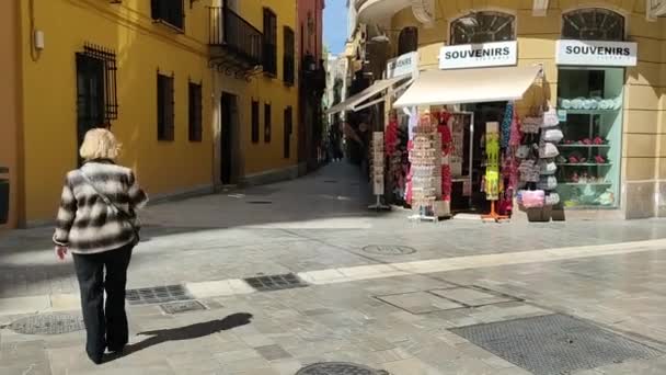 ブロンドの女性がマラガ市内中心部の道路を横断している スペインの街にあるお土産屋さん マラガの通りを歩く人々 スペインの都市都市の光沢のある通りのタイル — ストック動画