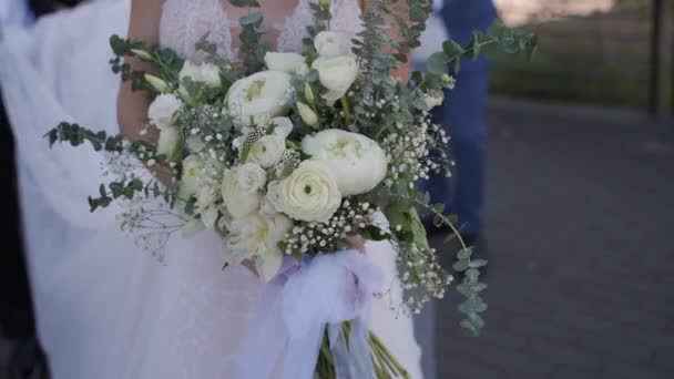 白いウェディングドレスを着た若い花嫁の手には長い尾があり 昼間に屋外を歩く手には花の花束があります — ストック動画