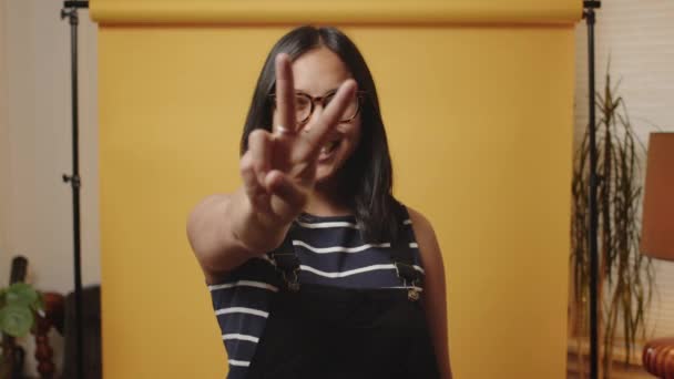 Happy Young Asian Woman Holds Peace Sign Camera Yellow Studio — стокове відео