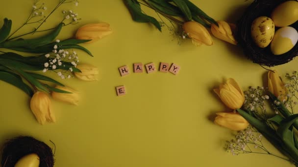 Top view of hands putting together scrabble letters as Happy Easter in the frame yellow eggs along with yellow tulips on yellow background.