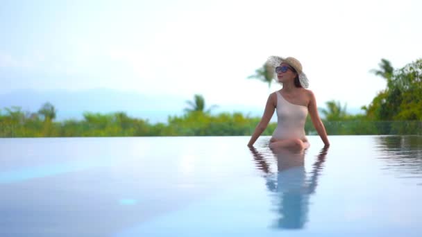 一个年轻的女人坐在一个无边无际的游泳池的浅端 伸出手来调整她的太阳镜 标题空间 — 图库视频影像