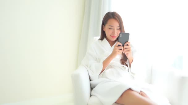 Happy Asian Woman Bathrobe Sitting Window Checking Messages Mobile Phone — Stockvideo