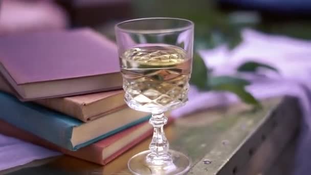 カラフルな本や木のテーブルの上に白い布の上に葉のスタックの横に白いワインのダイヤモンドスタイルのガラスの後方ボケショット このワインのグラスは花嫁と新郎のためです — ストック動画
