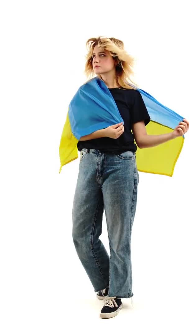 垂直ビデオ 若いブロンドの白人女性は暗い色の都市の服を着て 彼女の頭を回して立ってポーズをとり ウクライナの旗を示しています 白い背景のスタジオで撮影 — ストック動画