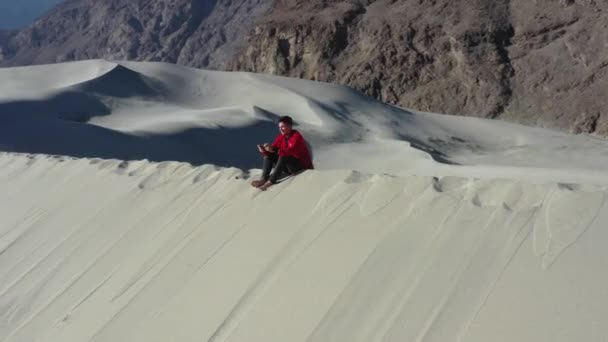 在一个阳光明媚的日子里 穿着红色夹克驾驶着无人驾驶飞机 坐在冰雪覆盖的沙漠中的沙丘上 远处是高山 — 图库视频影像