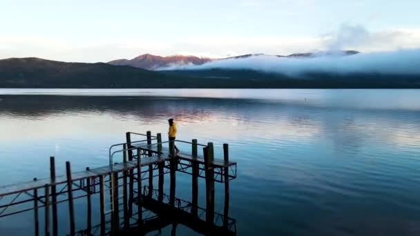女性は日没時に湖の木製の桟橋を歩く ニュージーランドの山々の美しい景色を楽しむアヒルの池を歩く黄色のジャケットの女性ドローンが飛んでいる間 — ストック動画