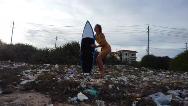 プラスチックでいっぱいのビーチをサーフボードで歩く若いサーファーの女の子の追跡ショット 環境汚染の概念 — ストック動画