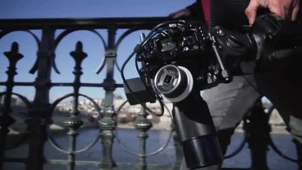 A rendező egy korláton sétál a Moldva-töltésen egy állványon lévő kamerával. Közelkép egy kamerát tartó kézről.