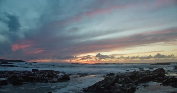 色彩斑斓的落日天空俯瞰着浪花在岩石上翻滚 这是在英国康沃尔小火海海滨的潮汐池中造成的 固定射击 — 图库视频影像