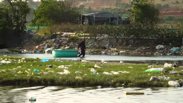 彼の神託船を持っている男が浅い川を歩いている 近くの川や土地はプラスチックで汚染されています — ストック動画