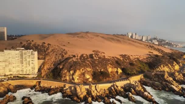 海の波の隣の険しい丘の中腹にあるオレンジ色の砂丘のうち 空中のドリー 曇りの日にコンコンアパートメンツとリゾート — ストック動画