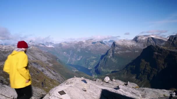 黄色のジャケットと赤い帽子を持つ男性ハイカーは 美しい景色を楽しむために山の崖の端まで歩いています ノルウェーの山岳地帯 ガイランガー フィヨルド — ストック動画