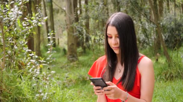 森の中に立っている間 美しい若いラテン系の女性がスマートフォンで閲覧しています 長いブルネット髪美しい赤いドレス — ストック動画