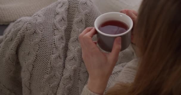 女孩坐在沙发上 裹着舒适的衣服和毛毯 一边喝着热咖啡 一边大放异彩 — 图库视频影像