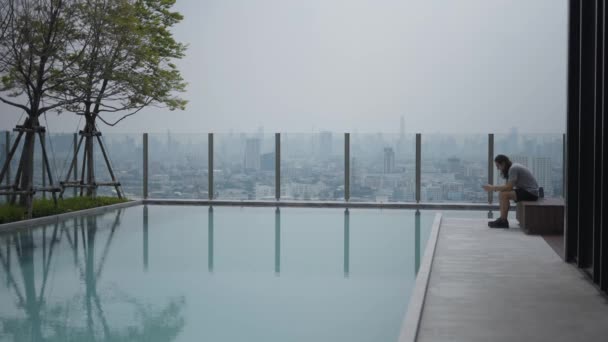 Lone Guy Using Smartphone Sitting Poolside Lounge Cityscape Background 곡괭이 — 비디오