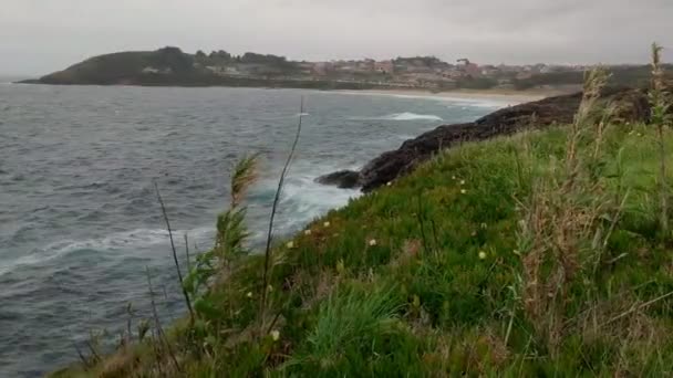 風の強い曇りの日に海の崖の上の植物波がクラッシュし 背景にある村 暗い嵐の雲 視点は右に回って撮影しました ポンテベドラ ガリシア スペイン — ストック動画