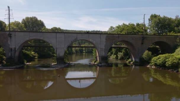 平らな茶色の川の水に完全に反映する列車の橋の下の歩道に立っている若い男 低空飛ぶショットを通して — ストック動画