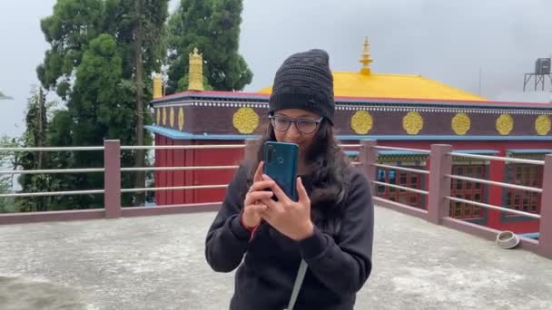 インドの冬の朝にスマートフォンの携帯電話を使用してスカーフを着て修道院の複合体の外に立っているインドの女の子 — ストック動画