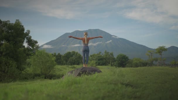 スポーティブロンド女性でヨガレギンス持ち上げ腕に上へ敬礼ポーズ 火山アグンで背景 — ストック動画