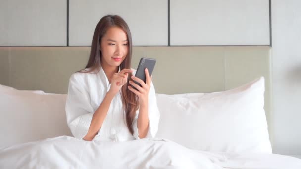 入浴後は一本指で携帯電話の画面に触れるアジア人女性 ホテルの部屋にあるバスローブのキングサイズベッドに座っている間 スマートフォンを使用して笑顔の女の子の肖像画 — ストック動画