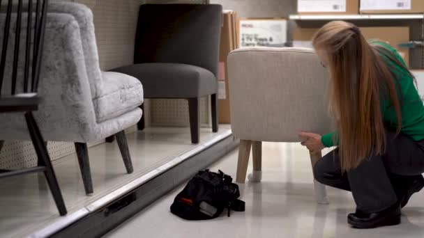 Squatting Blonde Woman Scrutinizes Upholstered Modern Looking Chair Store Display — Stock Video