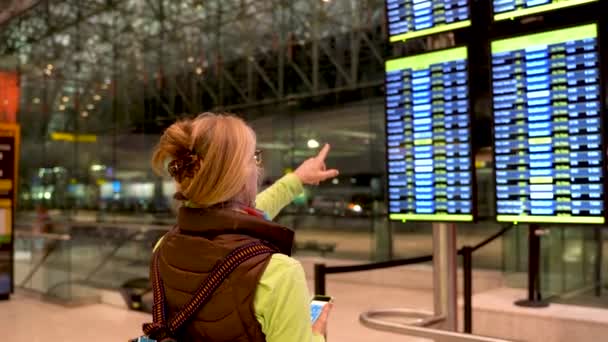 Blond Woman Points Flight Information Display While Holding Her Smartphone — Stockvideo