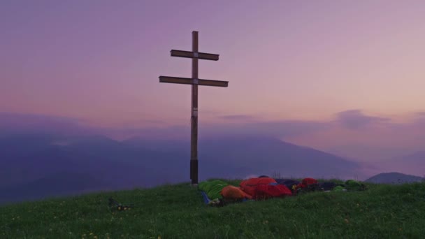 Group of people wild camping on the mountain peak with cross and green meadow. People in sleeping bags bivouacking in the nature before sunrise
