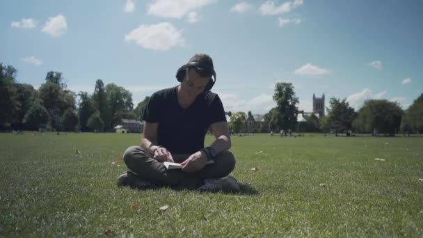 Young White Man Seated Grass Park Reading Book Sunny Day — ストック動画