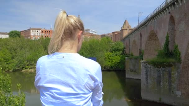 Saçları Arkadan Bağlanmış Sarışın Bir Kadın Nehrin Önünde Akıllı Telefonunu — Stok video