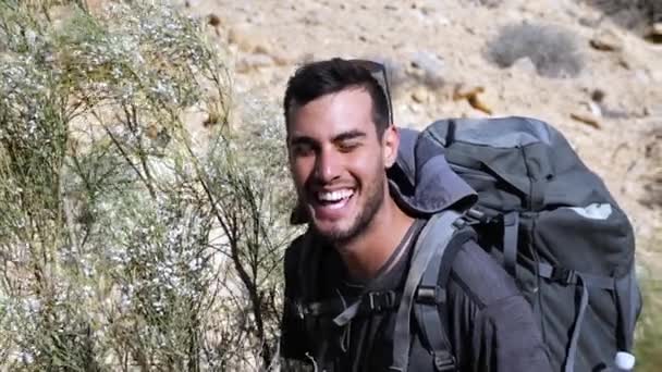 イスラエルのラモンクレータートレイルでブッシュを植えるために大きなリュックのうなずきの頭と笑顔を身に着けている大人の男性ハイカー スローモーション — ストック動画