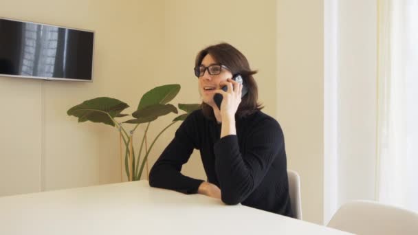 Young Man Glasses Smiles Talks His Cell Phone Sitting Table — Αρχείο Βίντεο