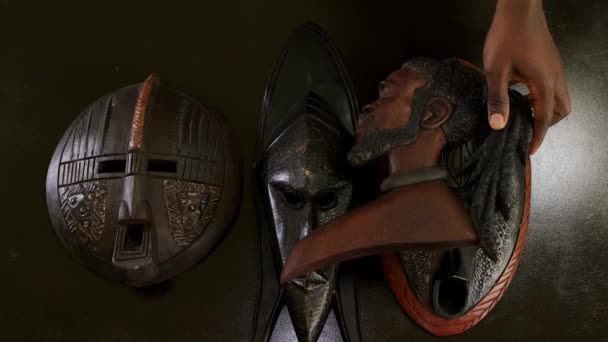他の木製のアフリカのマスクの上に男性と女性の木製のアフリカのマスクを積み重ねるアフリカ系アメリカ人の男性の手のトップダウンショット — ストック動画