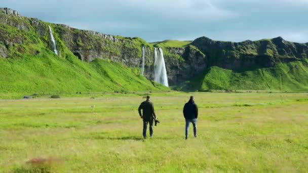 Two Tourists Walking Seljalandsfoss Gljufrabui Waterfalls Beautiful Field Grass Sunny — Video Stock