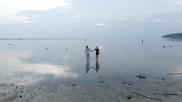 異人種間の旅行者のカップルは 日没時に島のビーチの浅い海の水から歩いて手を握っています 空中回転 — ストック動画