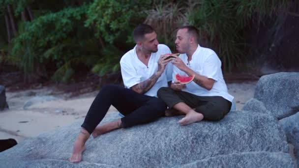 Happy Caucasian Gay Male Couple Eating Watermelon Sitting Rock Tropical — Stockvideo