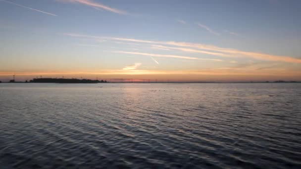 日の出時の湖の概要 光の風が水の中に素敵な波状の効果を引き起こします 背景には島と風車があります — ストック動画