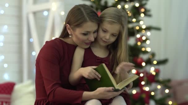 前面有一个小女孩和她的妈妈 他们正在一起看书 在背景上 我们看到灯光和圣诞树 它们都穿着红色的衣服 — 图库视频影像