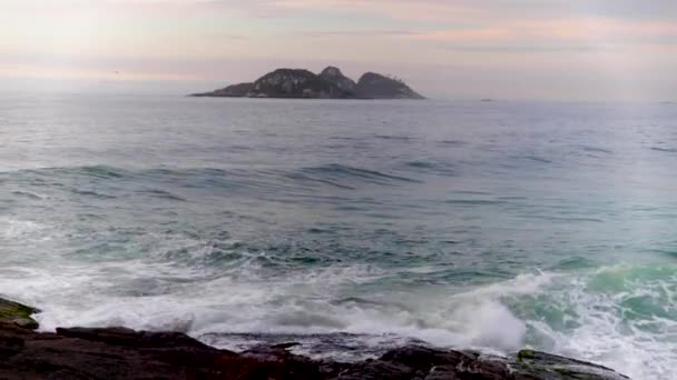 ジャネイロ海岸のすぐ外の水平線の美しい島岩のビーチに入ってくる前景波と背景にカラフルな日の出の空 — ストック動画